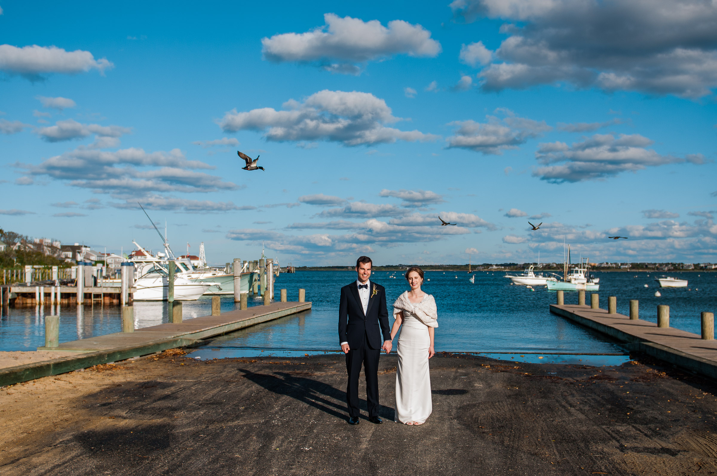 bride and groom holding hands in front of a boat ramp with a blue sky and puffy clouds in background on Cape Cod