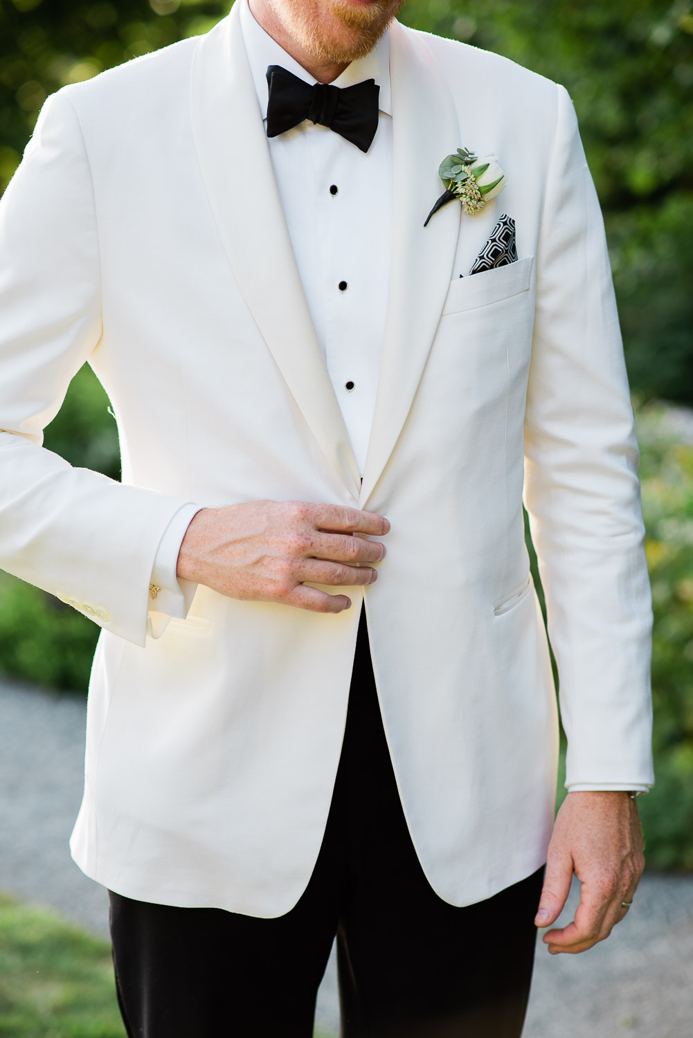 groom buttoning white tuxedo jacket with black pants and black bow tie at Blithewold Mansion