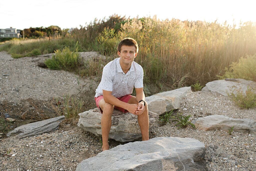 Sandy point senior portrait session with boy sitting on rocks in front of tall grass