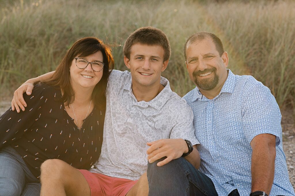 Sandy point portrait session with senior boy and Parents sitting on beach in front of tall grass in sunset