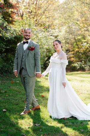 Bride and groom holding hands during fall wedding portraits at Wrights Mill Farm in Connecticut, with the groom in a green suit and the bride in an off-the-shoulder gown.