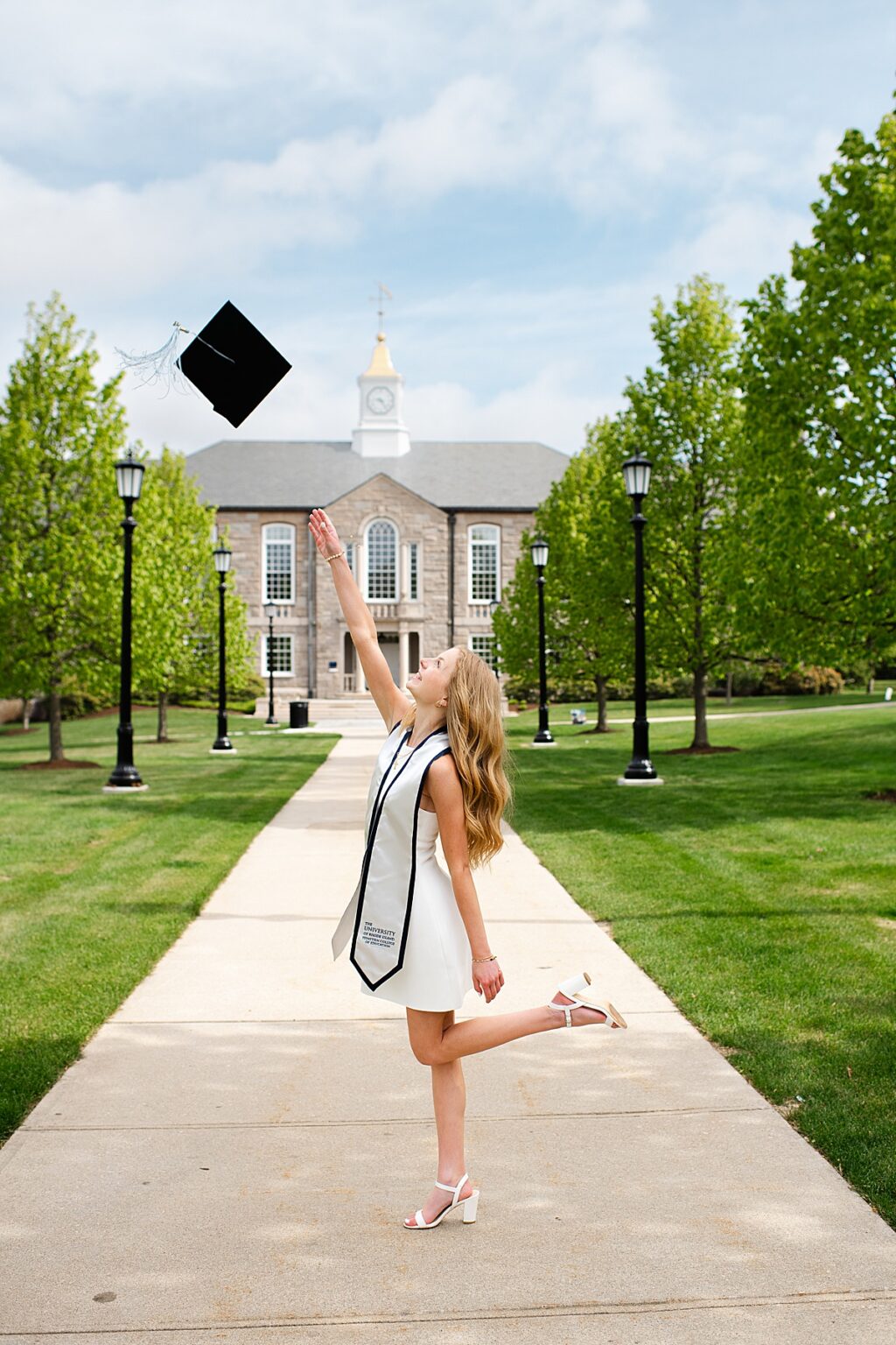 URI grad tossing cap in front of campus building lined with trees